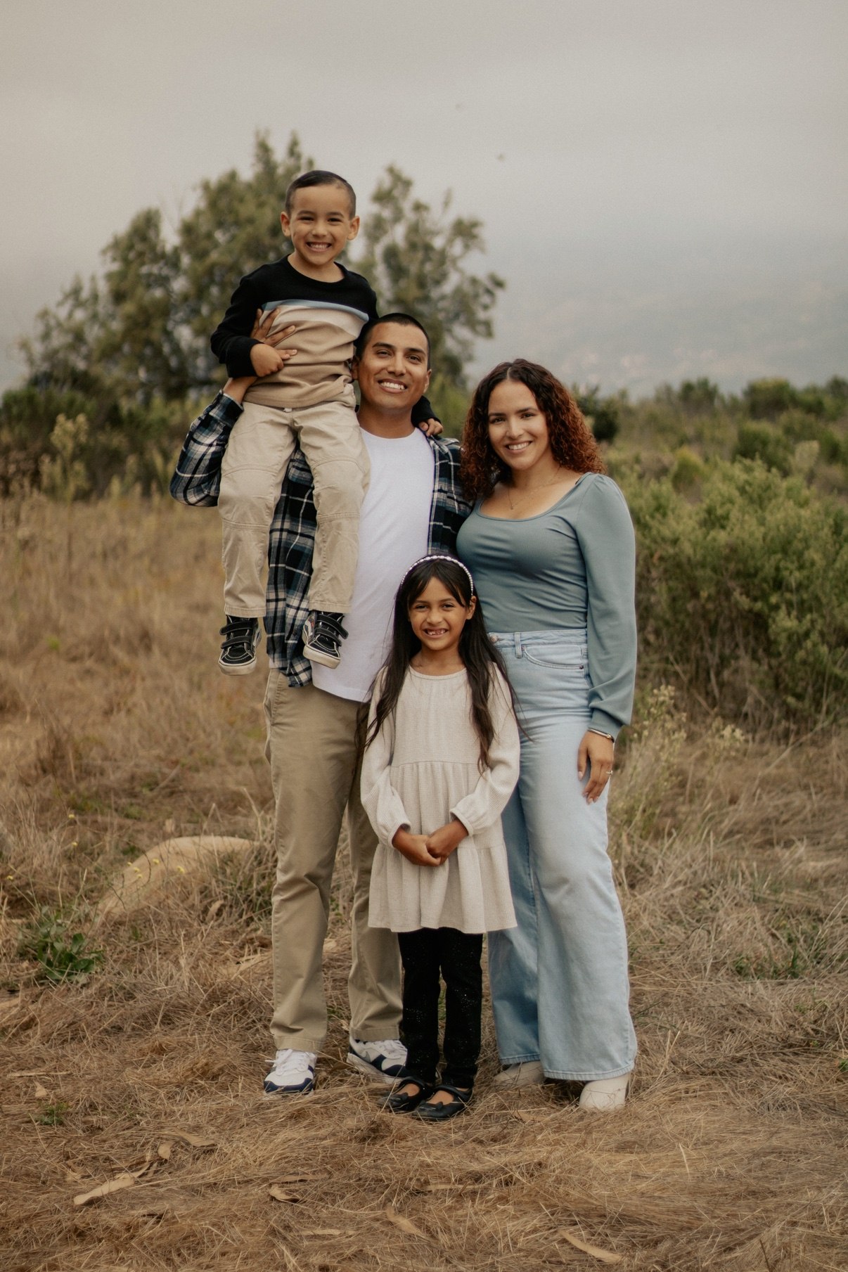 Family of four smiling in a field, young son riding on his father's shoulders
