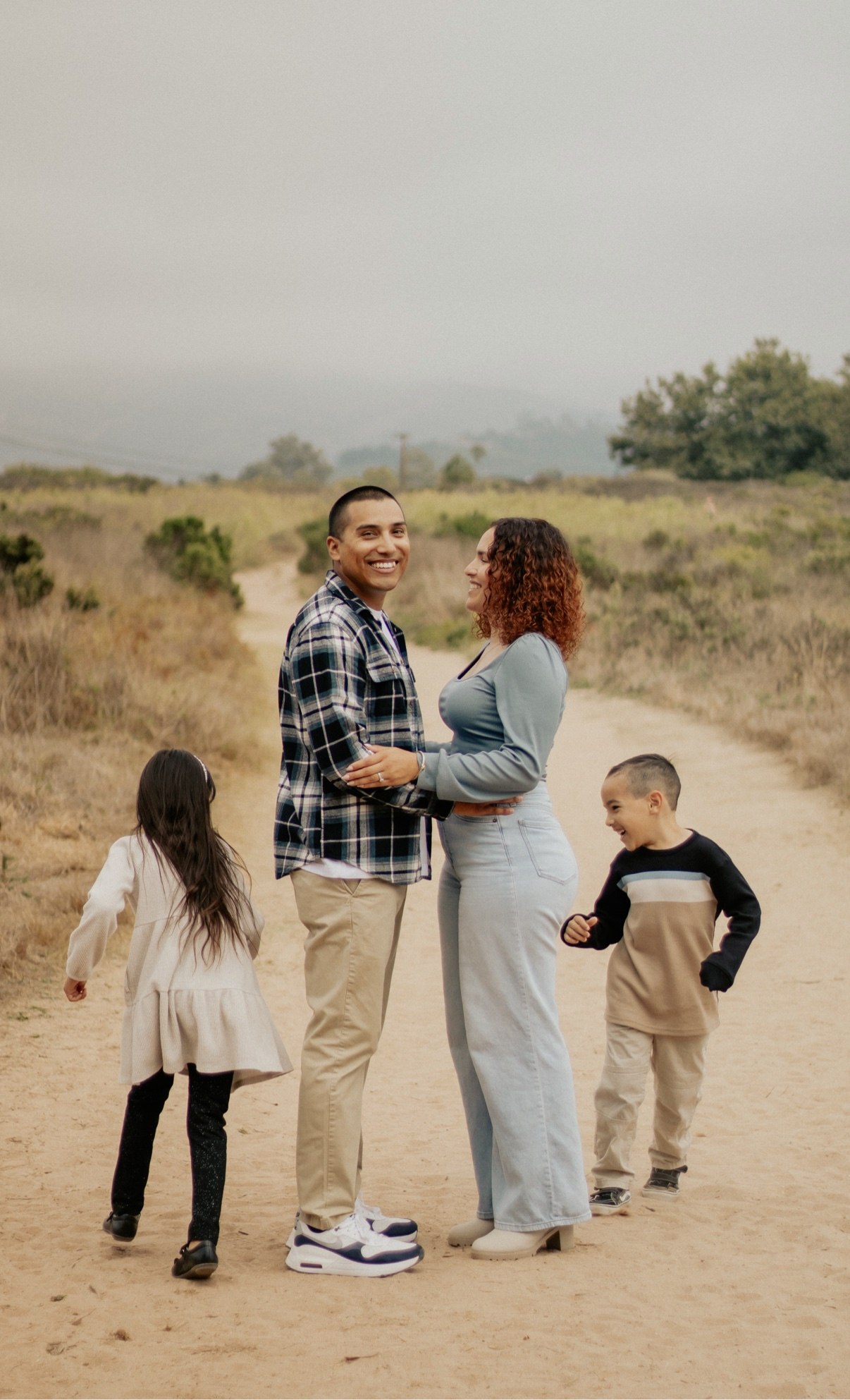 Parents holding hands and smiling on a dirt path while their two kids play around them