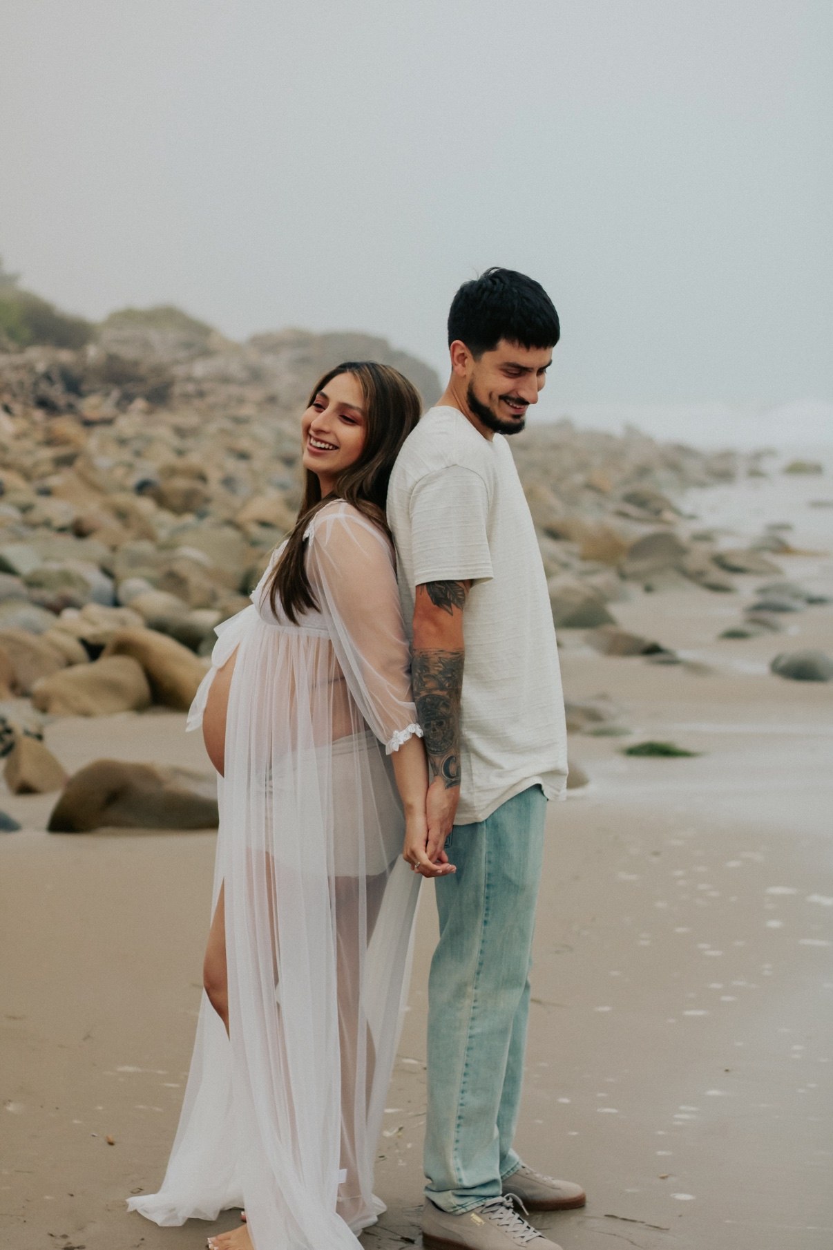 Expecting couple standing back to back on a rocky beach, holding hands and smiling