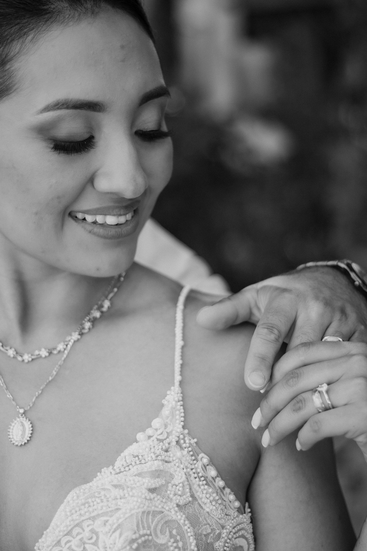 Black-and-white close-up of a smiling bride in a beaded lace gown, the groom's hand on her shoulder showing their rings