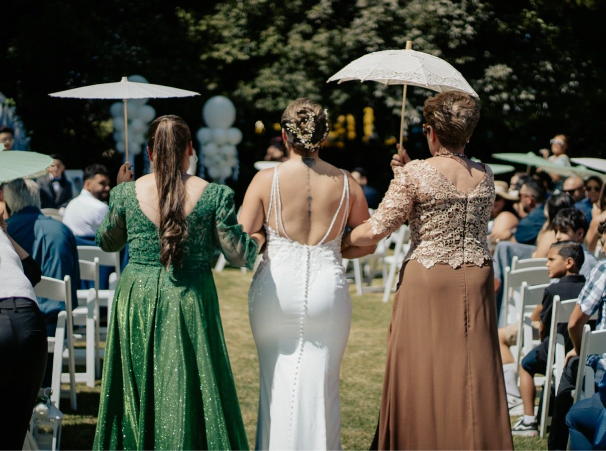 Bride seen from behind walking down the outdoor aisle between two women holding white parasols