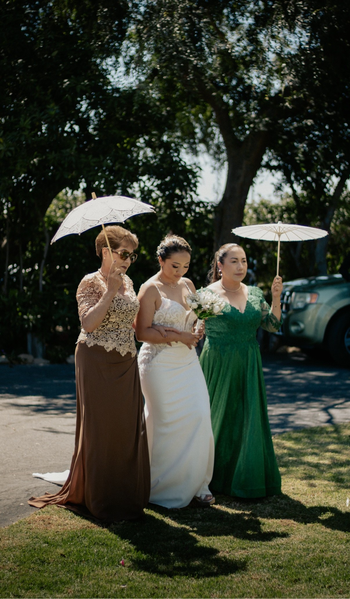 Bride holding a white bouquet standing between two women holding lace parasols under the trees