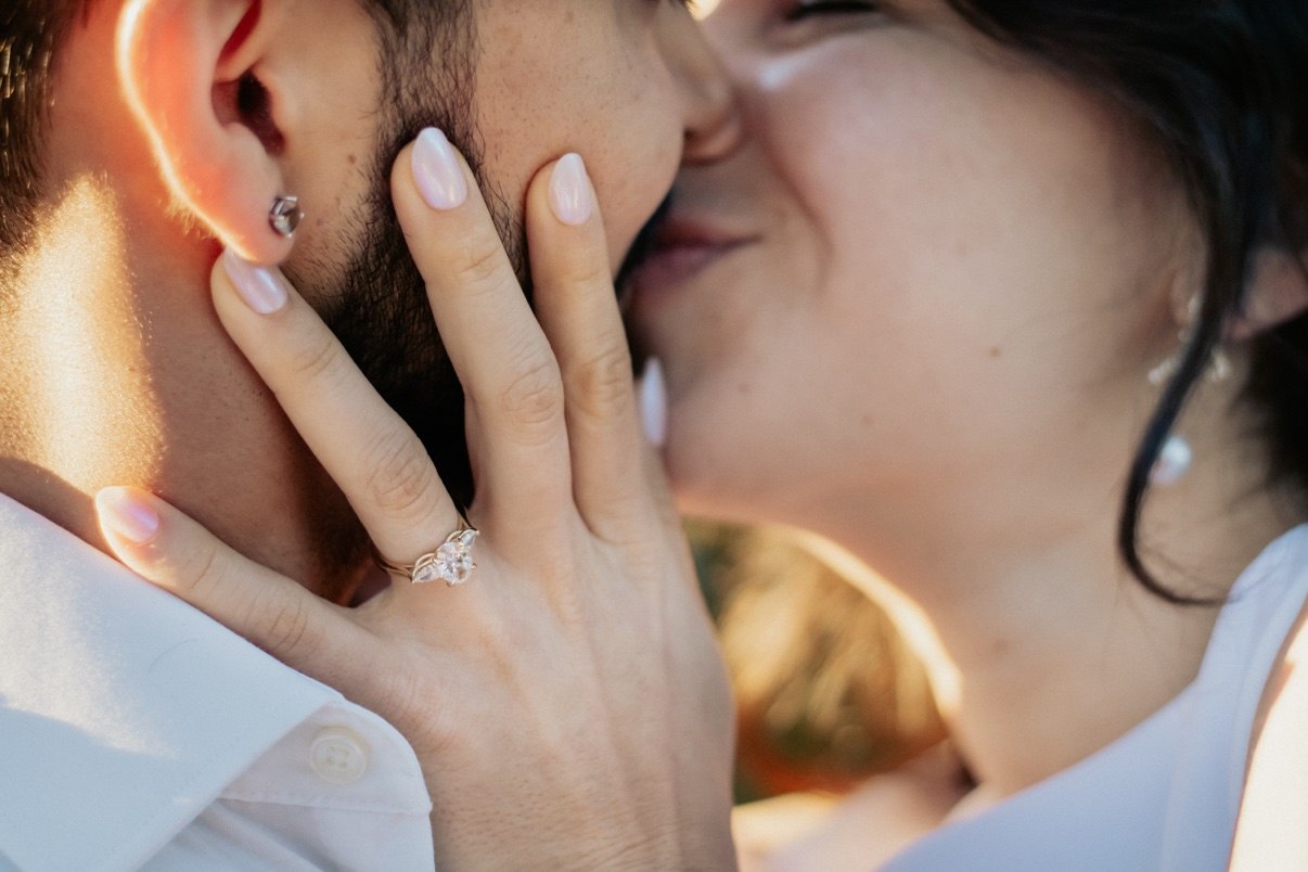 Close-up of a woman's hand with a diamond engagement ring cupping her partner's face as she kisses him