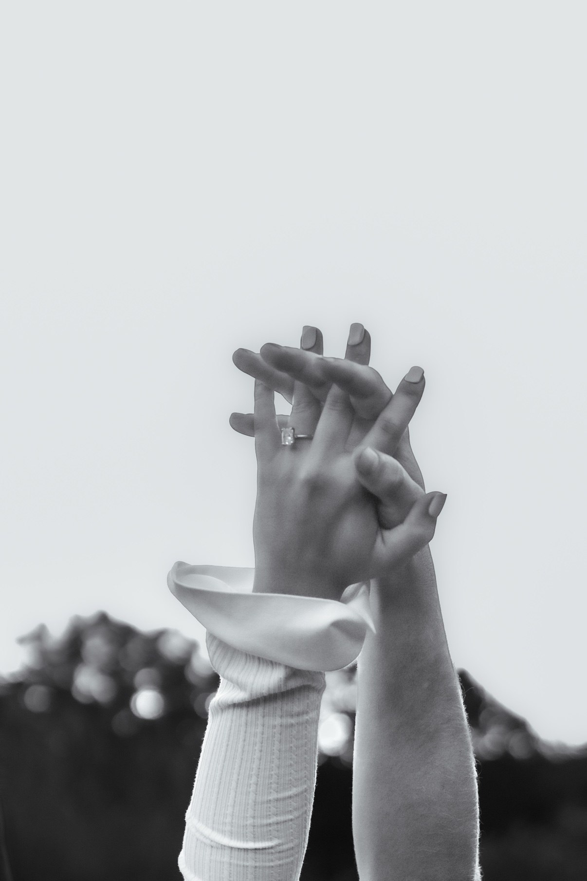 Black-and-white photo of a couple's hands intertwined against the sky, an emerald-cut engagement ring on her finger