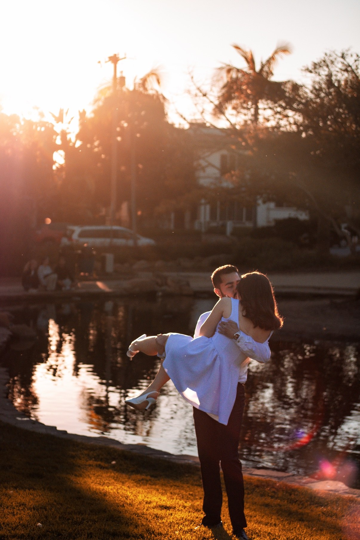 Man lifting and carrying his partner in a white dress beside a pond at sunset