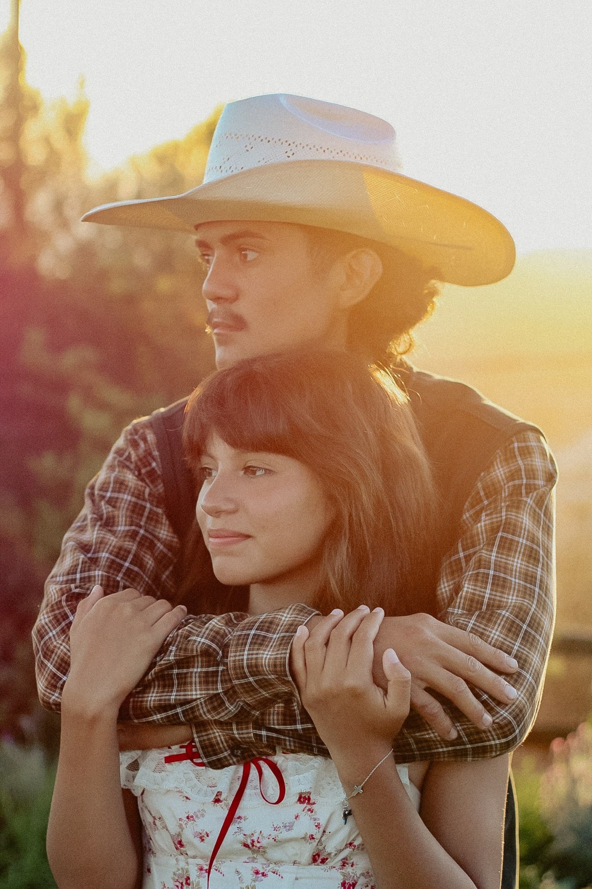 Man in a cowboy hat and plaid shirt embracing his partner in a floral dress at sunset