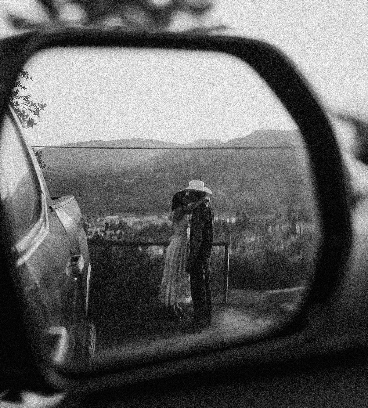 Black-and-white photo of a couple kissing, reflected in a car's side mirror with hills behind them